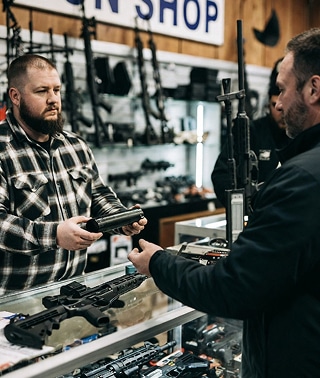 gun shop dealer works with customer from behind glass counter and is explaining differences in various firearms